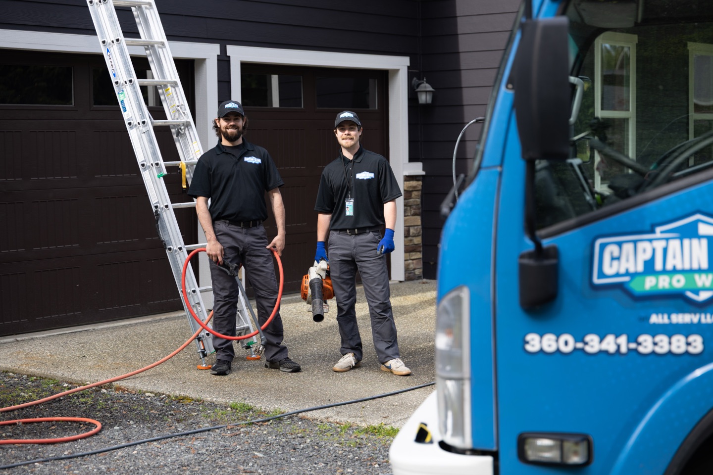 Two Captain Clean Pro Wash technicians with a hose and a leaf blower at a client's garage