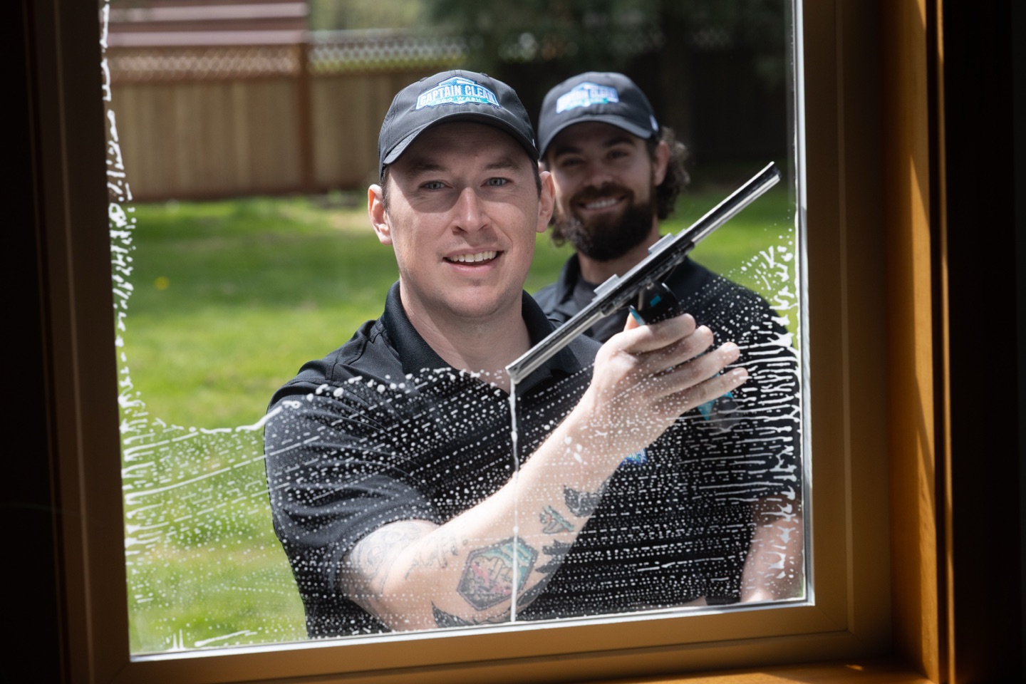 A Captain Clean Pro Wash technician squeegees soapy glass as a teammate looks on, seen from inside