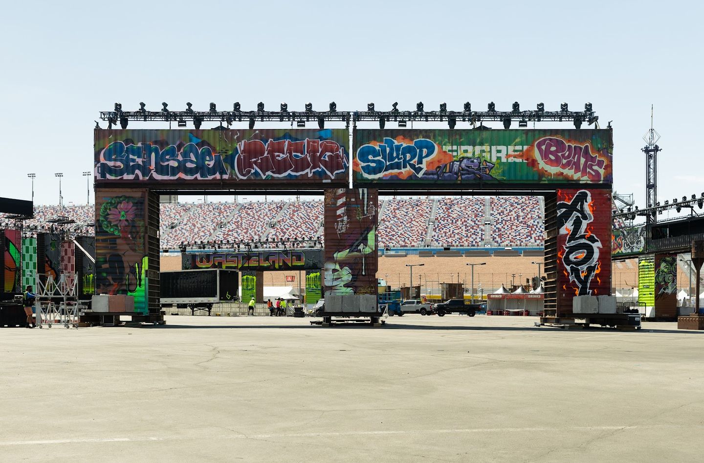 A gateway of stacked containers covered in painted lettering, the speedway grandstands behind